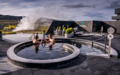 A couple relaxes in one of Krauma Spa’s geothermal pools, toasting with drinks as steam rises in the background. The modern outdoor spa, surrounded by Iceland’s scenic countryside, features sleek black stone pools with natural hot spring water from Deildartunguhver. A sign indicates the pool’s temperature of 40-41°C (104-106°F), while lounge chairs and greenery enhance the tranquil atmosphere.