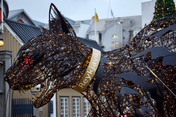 A large, illuminated wire-frame statue of the Icelandic Christmas Cat (Jólakötturinn) stands in a downtown area of Reykjavik. The cat is dark-colored with thousands of tiny golden lights woven into its skeletal structure, giving it a glowing appearance. It has sharp teeth and claws, and a collar with "Reykjavik" written on it. Buildings with traditional Icelandic architecture are visible in the blurred background under a cloudy sky.