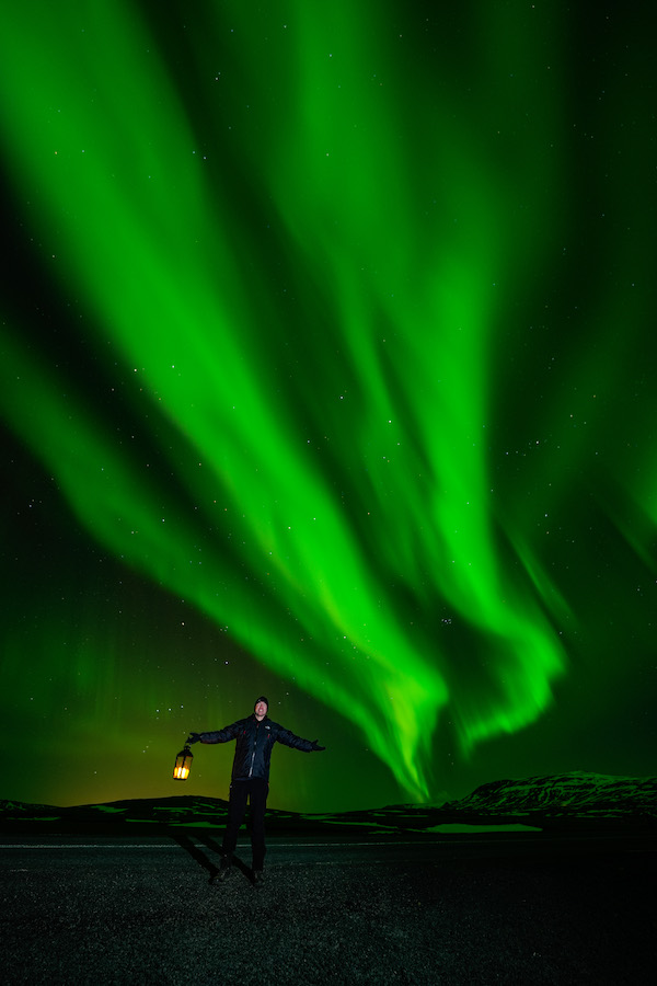 A man with a lamp stands in front of the northern lights in Iceland.