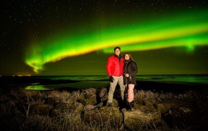 A man and woman standing together in Iceland with the northern lights in the background.