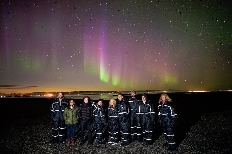 A group of people stands in front of the northern lights in Iceland.