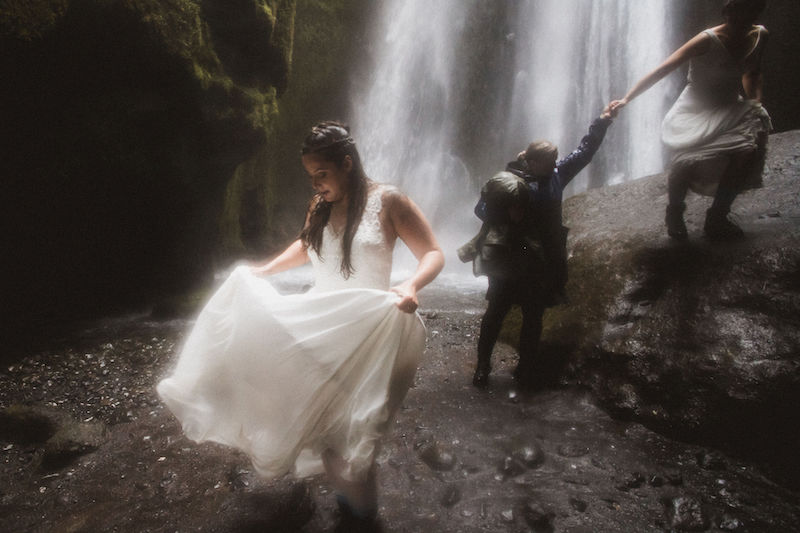 Two ladies in wedding dresses near an Icelandic waterfall.