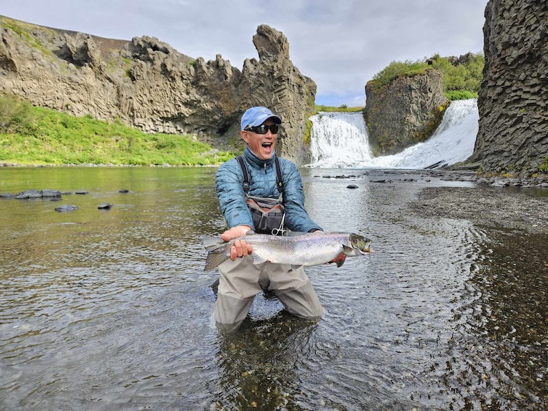 A man in a river holding a salmon at Fossá river in Thjorsárdalur valley in Iceland. Behind him is the waterfall Hjálparfoss
