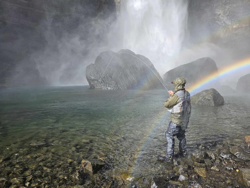 A man is fly-fishing in an Icelandic river with a rainbow and a waterfall next to him.