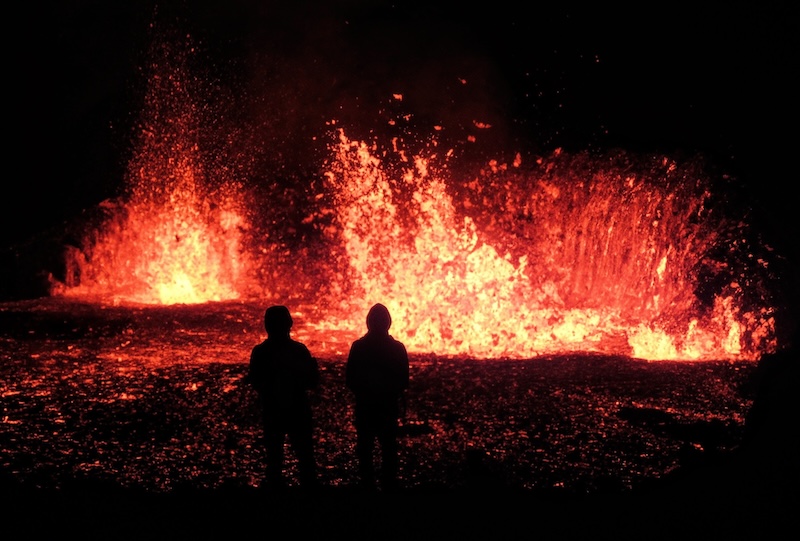 Two people stand in front of an erupting volcano in Iceland.