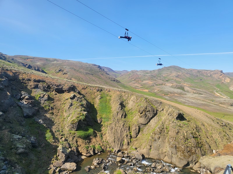 On a sunny day, two people over a canyon on a zipline in Iceland.
