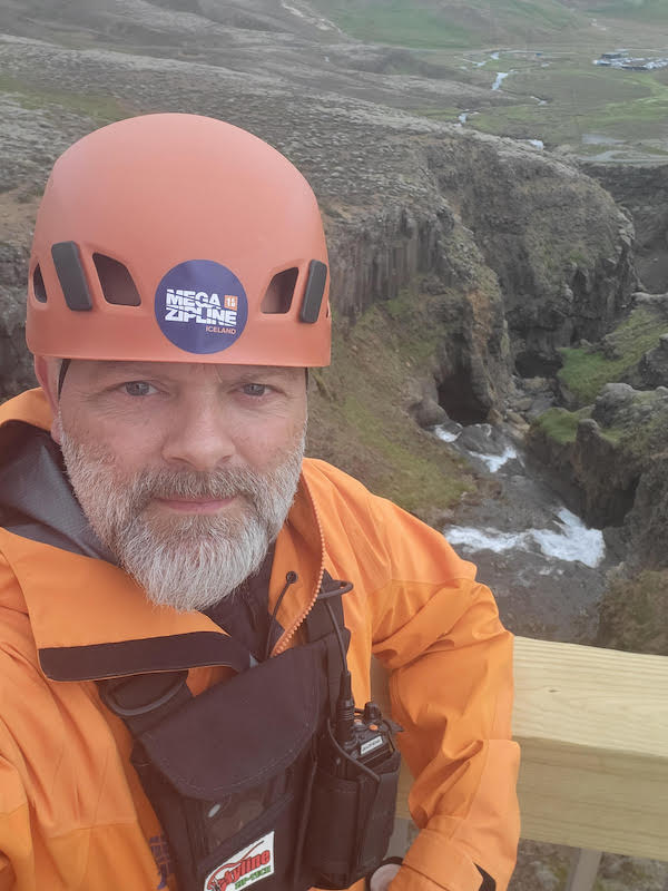 A man with an orange helmet stands before a canyon in Iceland.