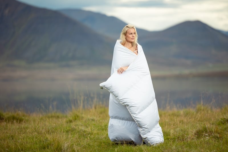 A blonde woman stands outside in Iceland, wrapped in an Icelandic eiderdown duvet.