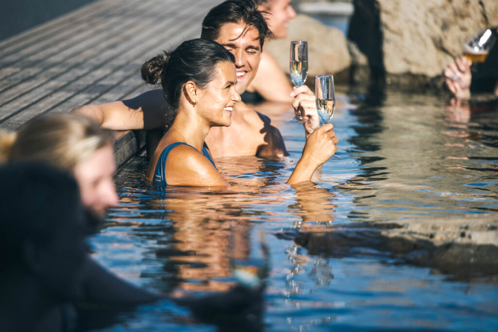 Friends toasting with champagne and beer in a geothermal pool at Hvammsvík Hot Springs in Hvalfjörður, Iceland.