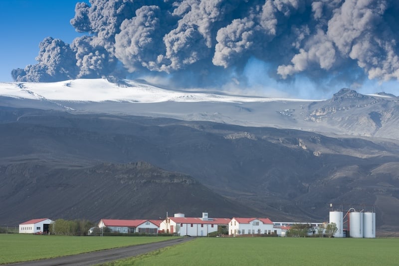 Eyjafjallajokull in Iceland erupting, ash plume against blue sky above the farm Thorvaldseyri, green fields in the foreground. Photo by Jóhann Helgason on Shutterstock.