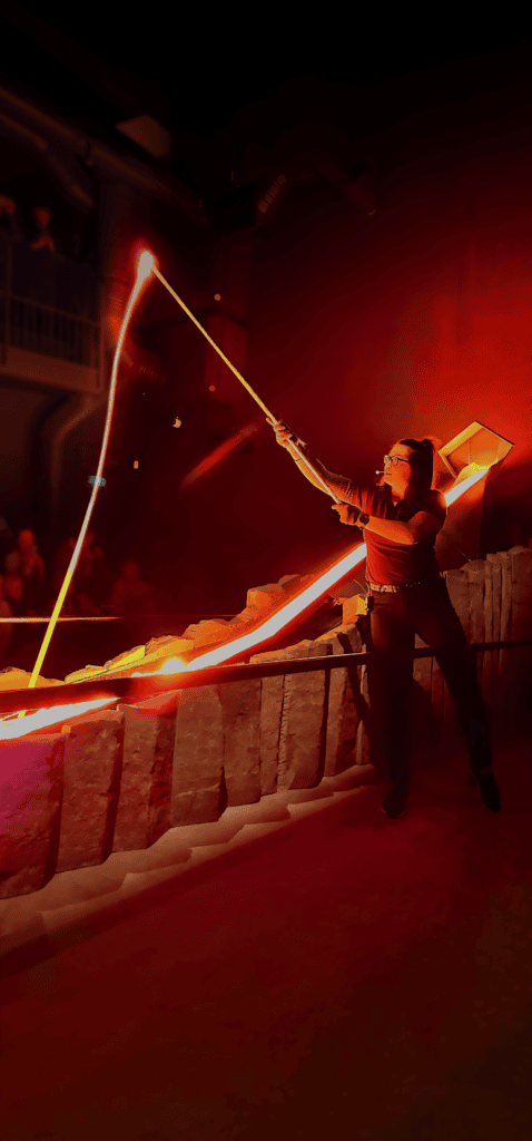 Lava Show performer lifting a long strand of glowing molten lava with a metal rod inside the dimly lit show room, with audience members visible in the background — a family-friendly indoor volcanic experience in Reykjavík and Vík, Iceland.