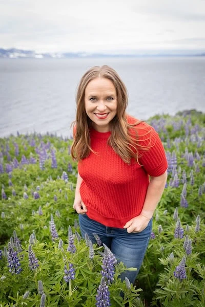 Portrait of Finnish author Satu Ramö standing in a blooming lupine field by the sea in Iceland, wearing a bright red sweater and smiling at the camera — a vibrant and natural setting reflecting her Nordic roots and literary connection to Iceland.