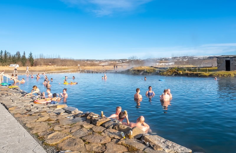 People relaxing in the geothermal waters of the Secret Lagoon, Iceland’s oldest hot spring located in Flúðir, surrounded by natural scenery and steam rising from the warm pool under a clear blue sky 
