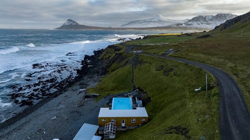 Remote geothermal pool Krossneslaug in the Westfjords of Iceland, perched on a grassy coastline with waves crashing against black volcanic rocks and snow-dusted mountains rising in the distance — a serene and dramatic destination for nature lovers seeking hot spring experiences in the wild.
