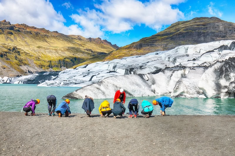 Group of hikers preparing for a glacier walk at Sólheimajökull, an outlet glacier in South Iceland, with dramatic ice formations, glacial lagoon, and surrounding volcanic mountains under a bright blue sky
