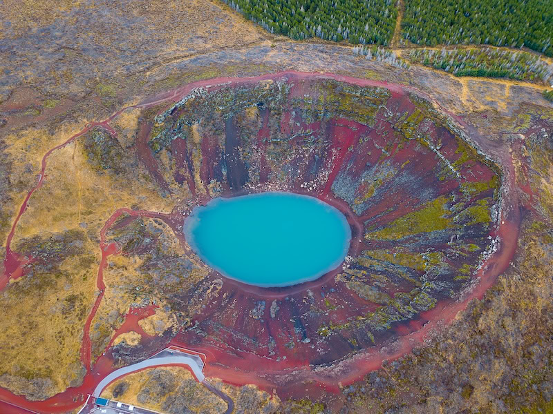 Aerial view of Kerið, a vibrant volcanic crater lake in South Iceland's Golden Circle, featuring striking red slopes and vivid turquoise water surrounded by moss-covered rock formations 
