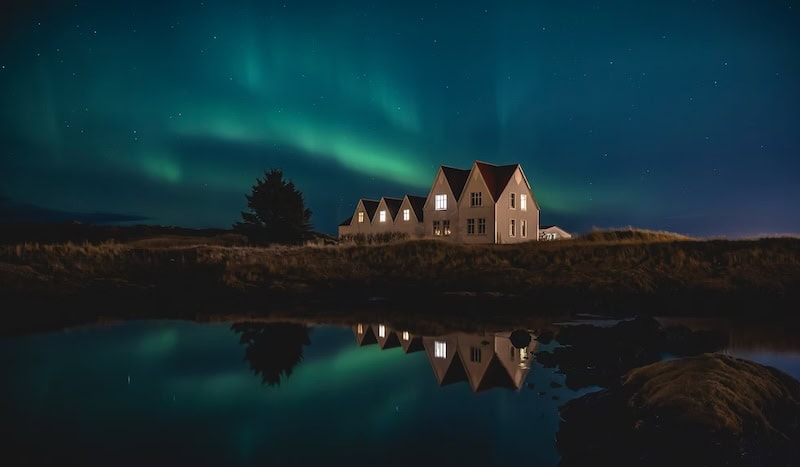 Traditional Icelandic houses reflected in a calm pond under the Northern Lights, with a starry sky glowing green and blue, photographed by Kévin Pagès in Iceland.