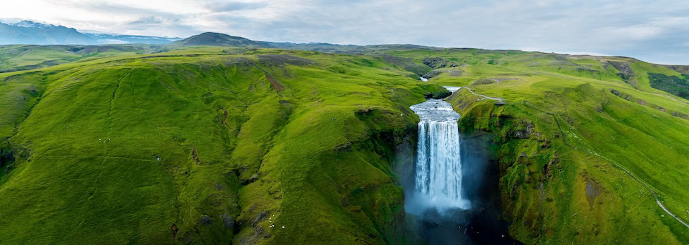 Aerial view of Skógafoss waterfall cascading over lush green cliffs in southern Iceland, a stunning place to cool off in Iceland during the summer, surrounded by dramatic highlands under a cloudy sky.