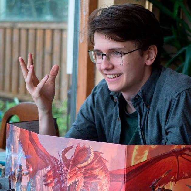 Magnús Guðrúnarson, a young man with glasses and dark hair, smiles and gestures while sitting behind a fantasy-themed game master screen featuring a red dragon. He appears to be running a tabletop role-playing game, seated indoors near a window.
