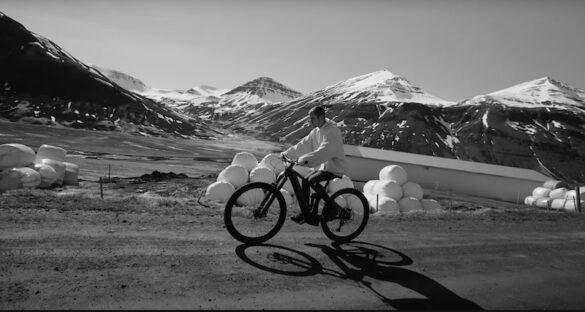 Justin Bieber in Iceland riding a bicycle through the scenic Fljót valley in North Iceland, with snow-capped mountains and hay bales in the background.