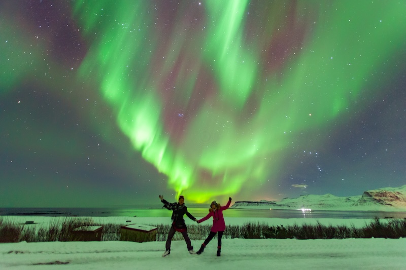 Two travelers enjoying the Northern Lights in Iceland during winter. Discover the best campervan deal for Iceland winter travel and explore the country’s stunning snowy landscapes at your own pace.