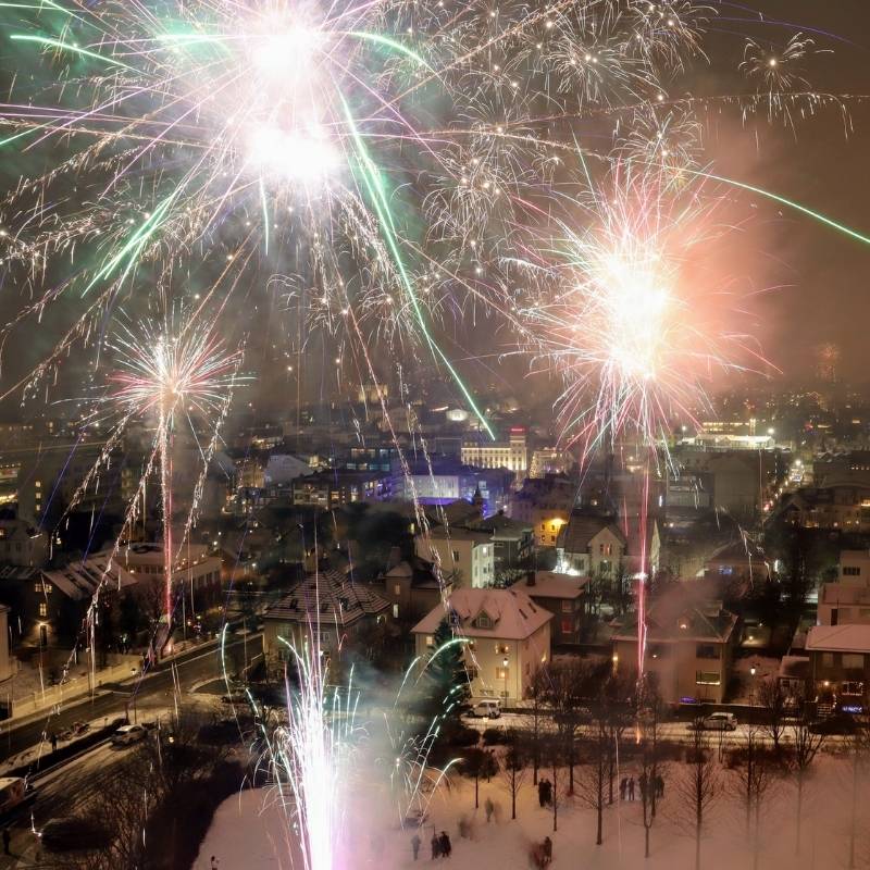 Fireworks bursting in green, pink and white over snow-covered rooftops in central Reykjavík at midnight on New Year's Eve, seen from above.