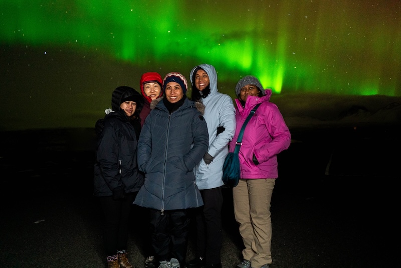 A group of people wearing outdoor clothes underneath the northern lights in Iceland.