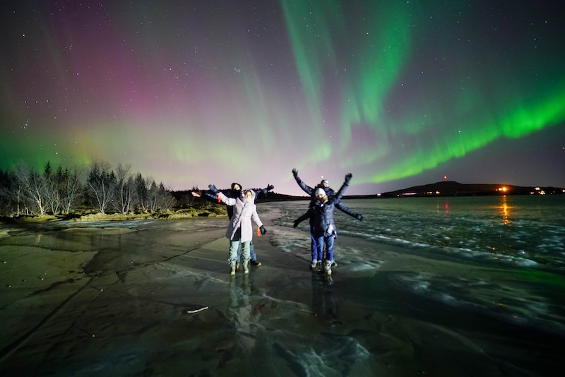A group of people wearing outdoor clothing on a frozen lake, dancing underneath the northern lights in Iceland.