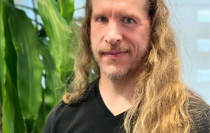 Portrait of Sigurjon Fridrik Gardarson, Emmy-winning compositing supervisor at Stormborn Studios, with long blonde hair, wearing a black shirt, standing in front of green indoor plants.