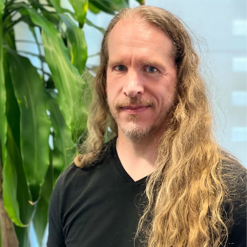 Portrait of Sigurjon Fridrik Gardarson, Emmy-winning compositing supervisor at Stormborn Studios, with long blonde hair, wearing a black shirt, standing in front of green indoor plants.
