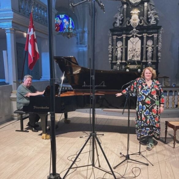 Soprano Hallveig Rúnarsdóttir and pianist Ulrich Stærk during a recording session in a grand church interior. Ulrich is seated at a Bösendorfer grand piano, while Hallveig stands beside a microphone wearing a colorful floral dress. The background features an ornate altar with classical sculptures and a stained-glass window, and the Danish flag hangs to the side. Recording equipment and microphones are visible in the foreground.