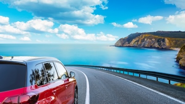 A red car drives along a scenic stretch of Iceland’s Ring Road, curving beside the ocean with dramatic cliffs and a bright blue sky in the background — a perfect moment which can be recreated by using the Stuck in Iceland Ring Road guide.