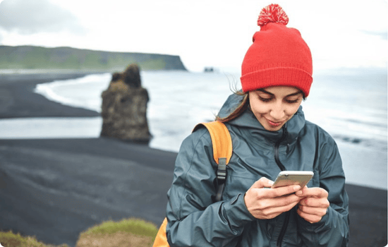 Traveler wearing a red beanie and backpack checks their phone while exploring a black sand beach on the south coast of Iceland.