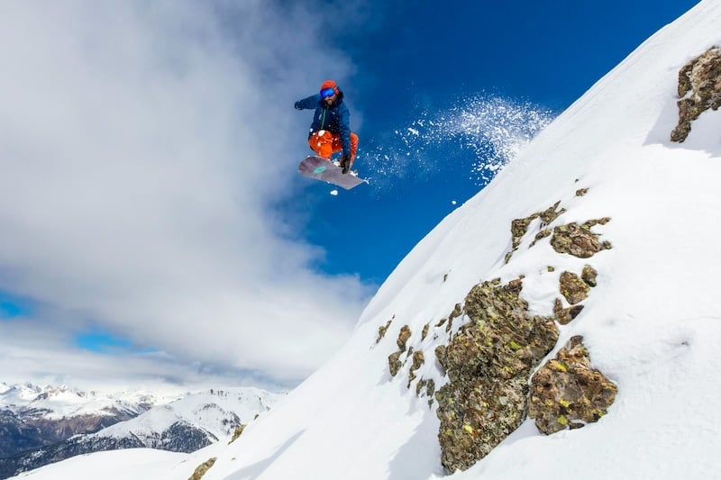 A snowboarder catches air while riding down a steep, snowy mountain slope in Iceland, with rugged rocks and stunning winter landscapes in the background.