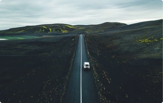Aerial view of a white campervan driving along a straight road through black volcanic landscapes in Iceland, with green hills in the distance.