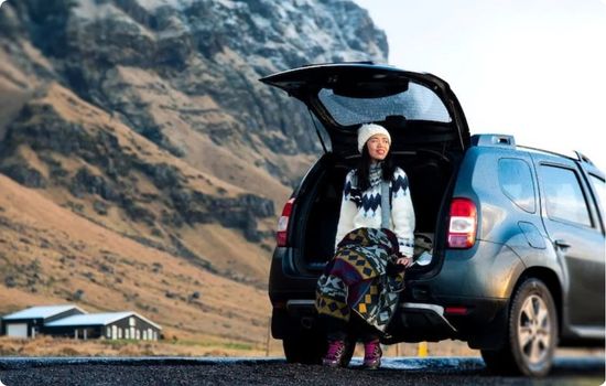 Woman sitting in the open trunk of a rental car during an Iceland road trip vacation, surrounded by dramatic mountain scenery and open countryside.