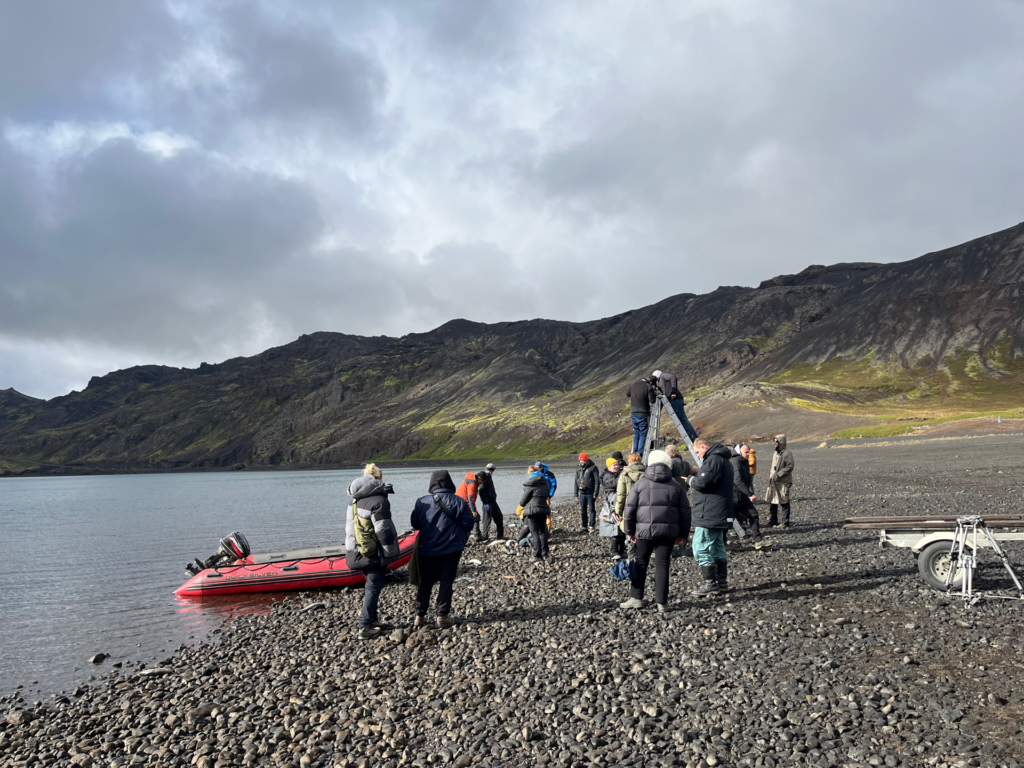 A film crew filming a scene from the movie The Fires.The crew is on a beach by a lake. A red boat is moored on the beach.
