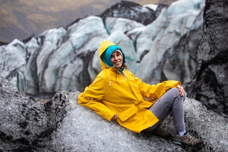 Female hiker in bright yellow waterproof jacket sitting on volcanic rock at Sólheimajökull glacier with blue-white ice formations and crevasses in background