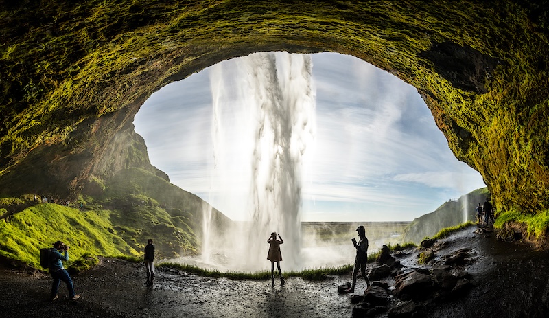 Seljalandsfoss waterfall in South Iceland viewed from behind the cascade with tourists silhouetted against moss-covered cave opening"
