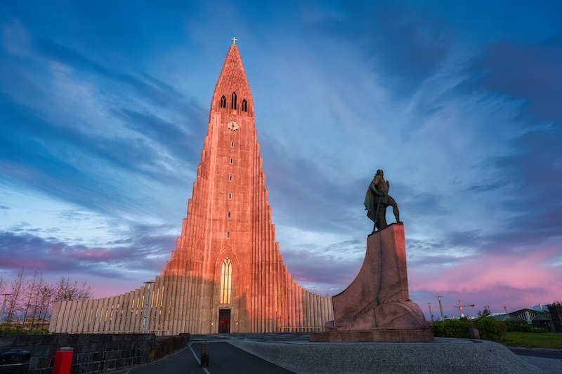 Long exposure photograph of Hallgrímskirkja church in Reykjavik at dusk, showing its distinctive white concrete tower and columnar facade illuminated in warm lighting, with the statue of Leif Erikson in the foreground and dramatic blue and pink twilight sky with motion-blurred clouds.