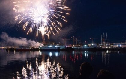 Fireworks exploding over Reykjavík's old harbour at night during Culture Night, with boats docked at the pier and colorful reflections shimmering across the dark water.