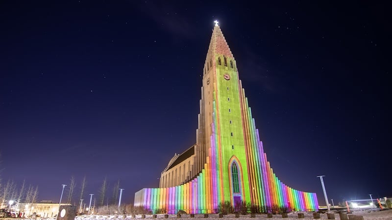Hallgrímskirkja church in Reykjavík illuminated with vibrant rainbow-colored light projections across its iconic stepped façade against a dark winter night sky, with a bright star shining above the tower's peak.