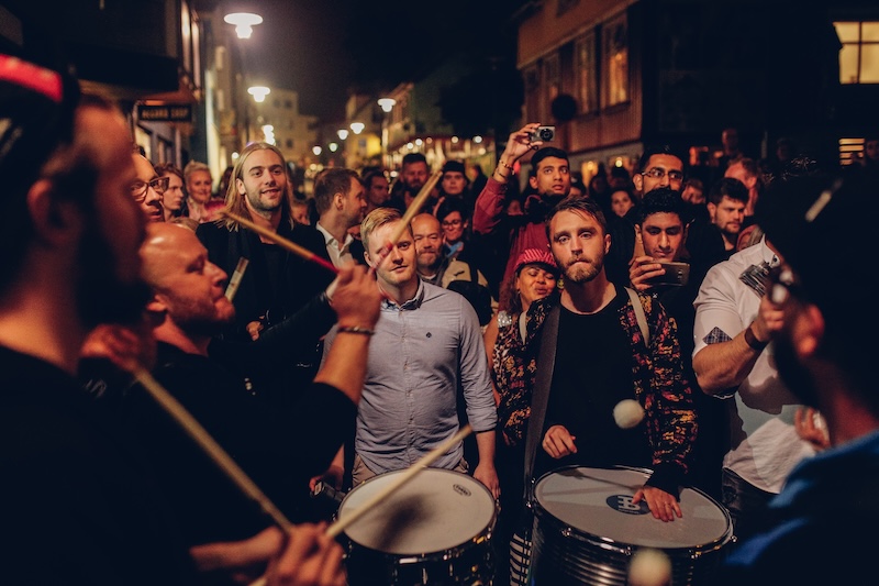 A crowd gathers on a Reykjavík street at night to watch a group of drummers performing. Several musicians play snare drums and hand drums while onlookers film with their phones, the scene lit by warm streetlights.