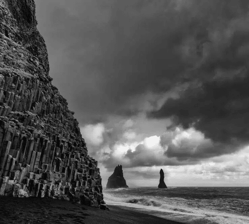 Reynisdrangar sea stacks rising from the Atlantic surf at Reynisfjara black sand beach, with dramatic basalt columns and stormy skies on Iceland's South Coast