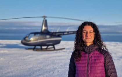 Helena Gallardo, customer service manager at Volcano Heli helicopter tour company in Iceland, standing on a glacier with a Robinson R66 turbine helicopter in the background on a clear winter day.