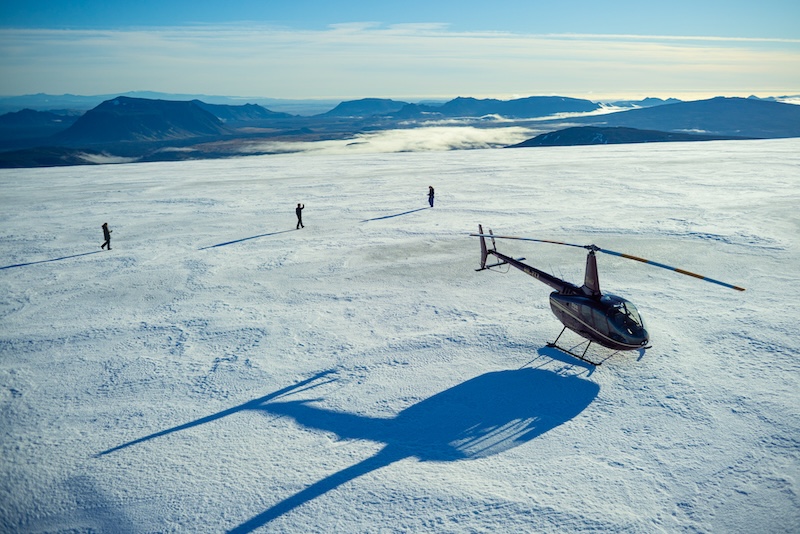 A Robinson R66 helicopter from Volcano Heli landed on a glacier in Iceland, with three passengers walking on the snow and panoramic views of Iceland's highlands and mountains in the background.