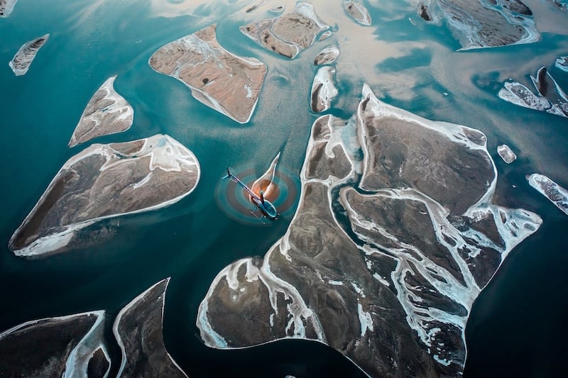 Aerial view of a Volcano Heli helicopter flying over a turquoise glacial lake with scattered sediment islands and glacial runoff patterns in Iceland's highlands.