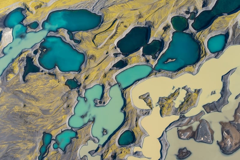 Aerial view of Iceland's highland interior showing vivid turquoise geothermal lakes surrounded by yellow-green moss, grey volcanic rock, and glacial sediment.