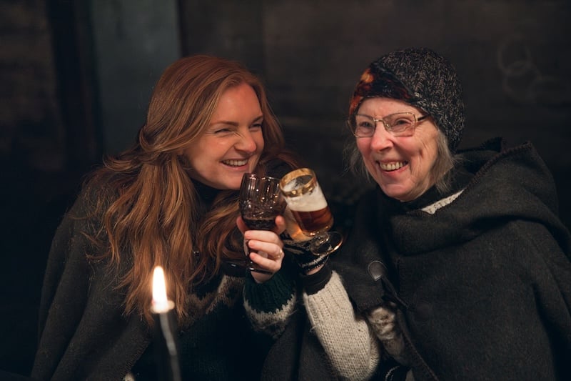 Two guests in Icelandic wool capes toasting with beer mugs by candlelight at Viking Story Night, Mr. Iceland farm, South Iceland