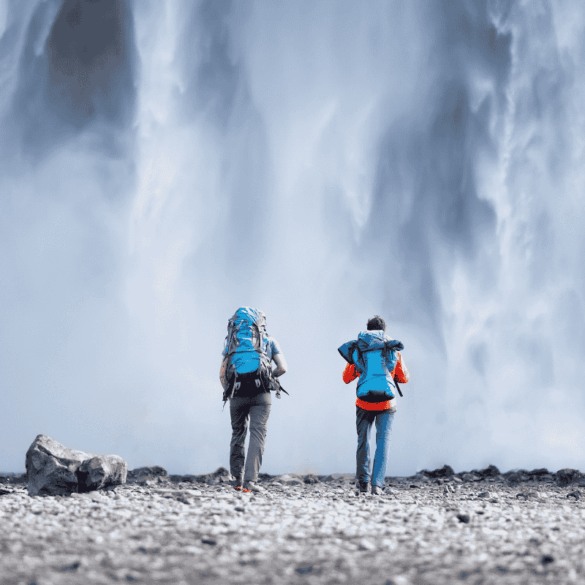 Two hikers with backpacks standing in front of a massive waterfall in Iceland — the kind of moment you can afford more of when you save on your 2026 Iceland vacation